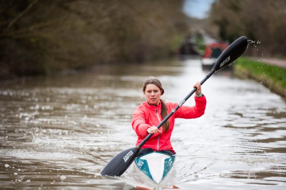 Person kayaking on a canal, surrounded by trees and a blurred background.