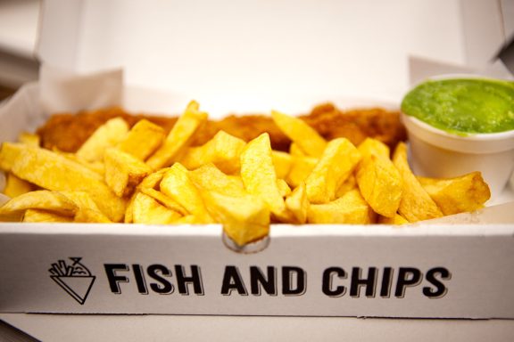 Fish and chips served in a takeaway box with a side of mushy peas.