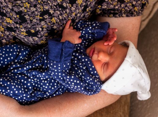 A person holding a sleeping newborn dressed in a blue polka dot outfit and white hat.