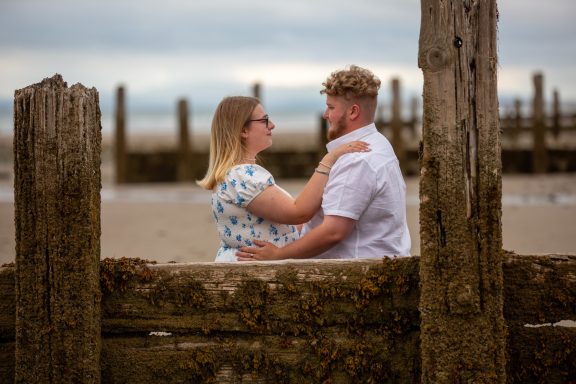 A couple embracing by the beach, framed by wooden posts, under a cloudy sky.