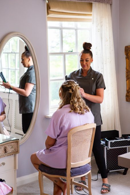 A stylist does hair in a bright room while another person sits in a chair, facing a mirror.