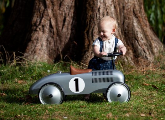 A young child plays with a vintage-style pedal car in a grassy outdoor setting.