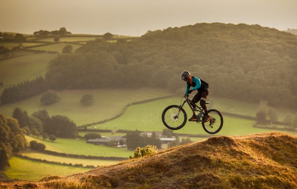 Mountain biker performing a jump over hilly terrain with a scenic landscape in the background.
