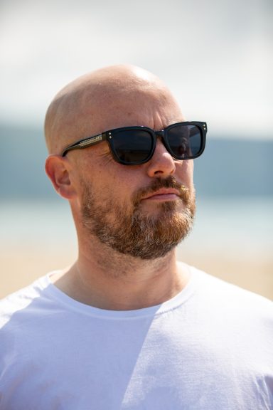 Bald man in sunglasses and a white t-shirt, looking thoughtfully into the distance outdoors.