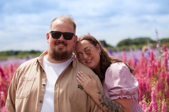 A smiling couple stands among vibrant pink flowers on a sunny day.