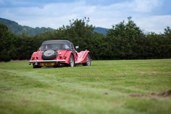 A red vintage car parked on a grassy field with trees in the background.