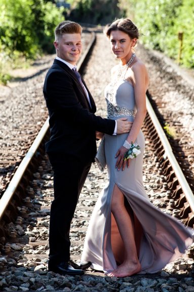 A couple in formal attire stands on a railway track, surrounded by greenery.