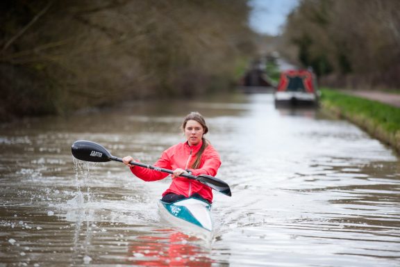 A woman kayaking on a calm canal with trees and a boat in the background.