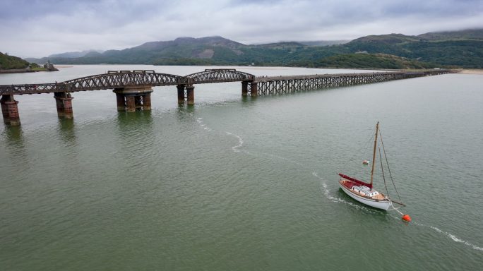 A small boat sailing near a long pier with mountains in the background.