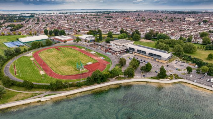 Aerial view of a sports facility with a track, buildings, and a waterfront nearby.