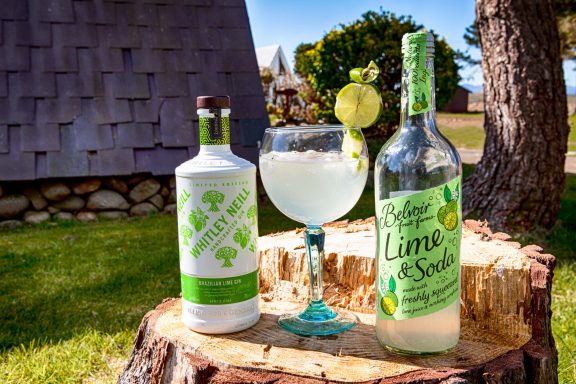 A glass of lime cocktail on a wooden stump, surrounded by spirit bottles.