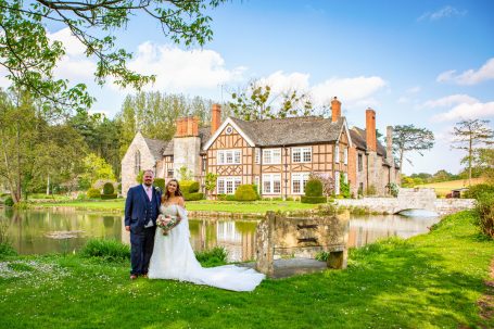 A bride and groom pose by a picturesque country house near a serene lake.
