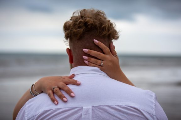A couple embraces on the beach, with a focus on their intertwined hands and rings.
