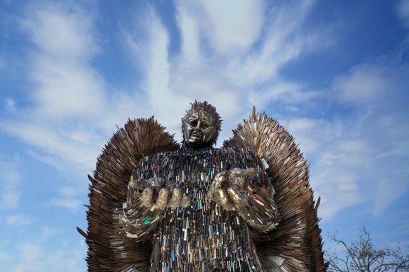 A large metal sculpture of a figure with wings, set against a blue sky with clouds.