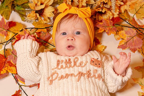 Baby wearing a yellow headband and sweater, surrounded by autumn leaves.