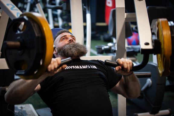 A man with a beard performs a bench press at a gym.