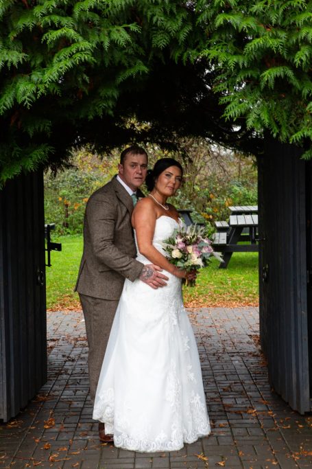 Bride and groom posed together under a leafy archway, holding a bouquet.
