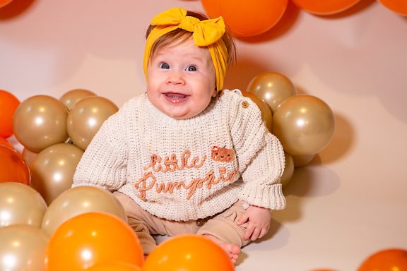Smiling baby in a cream sweater with a yellow bow, surrounded by orange and gold balloons.