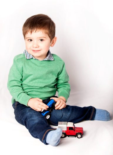 Young boy in a green sweater playing with toy cars on a white background.