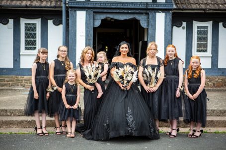 A bride in a black gown stands with eight bridesmaids in matching black dresses outside a house.