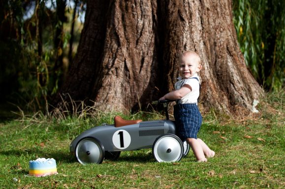 Toddler smiling while playing with a vintage toy car near a tree.
