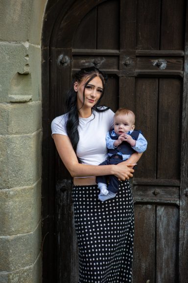 A woman holding a baby stands near an old wooden church door.