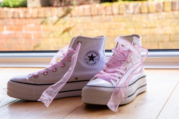 White high-top Converse trainers with pink laces and platform soles, set on a wooden floor.
